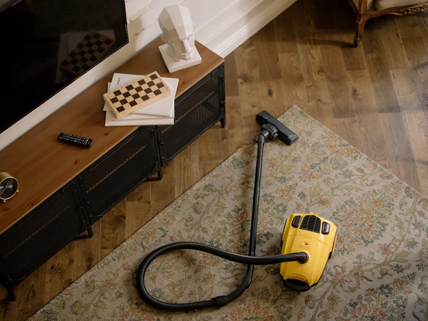 A person using a yellow and black vacuum cleaner to perform deep cleaning on a patterned area rug in a living room. The individual is placing a white cloth inside the vacuum's bag compartment, with th